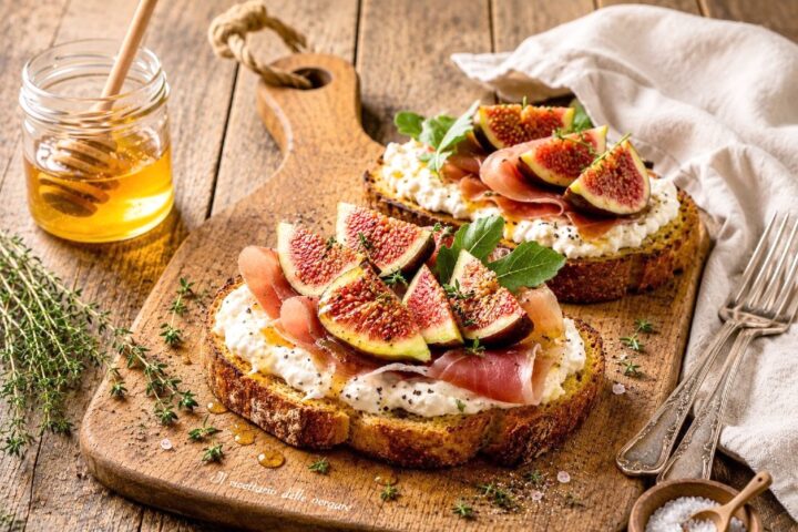Due bruschette con stracciatella, prosciutto crudo e fichi freschi a spicchi su un tagliere di legno.