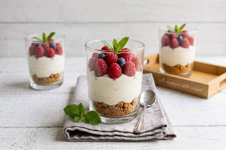 Tre bicchieri di cheesecake monoporzione con base di biscotti sbriciolati, mousse allo yogurt bianco e lamponi freschi in superficie.