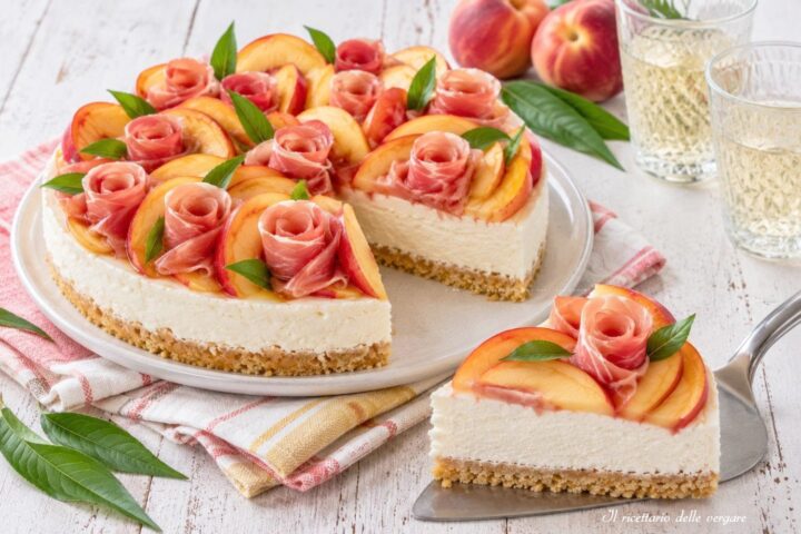 Cheesecake salata con base di biscotti salati, crema bianca, decorata con fettine di pesche fresche e rose di prosciutto crudo, con una fetta tagliata in primo piano.