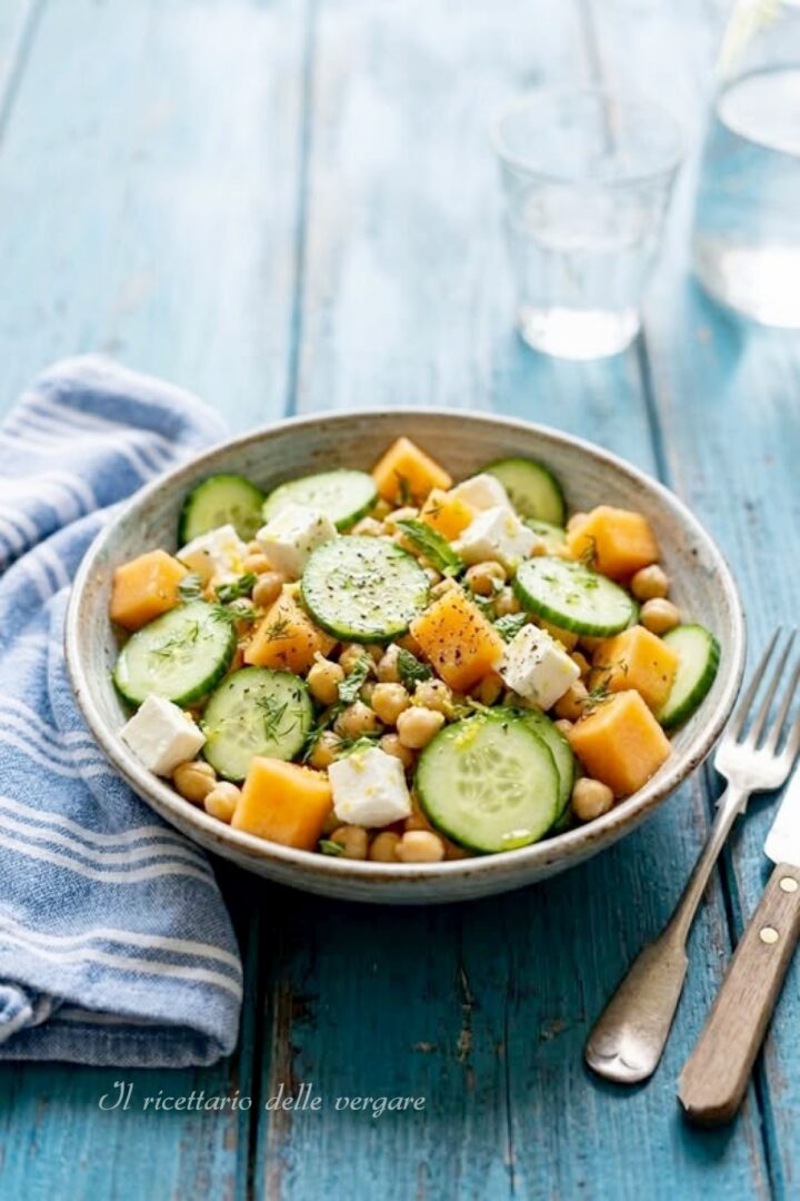 Inquadratura verticale di un'insalata di ceci con melone, feta e cetrioli servita in una ciotola su un tavolo di legno azzurro con posate