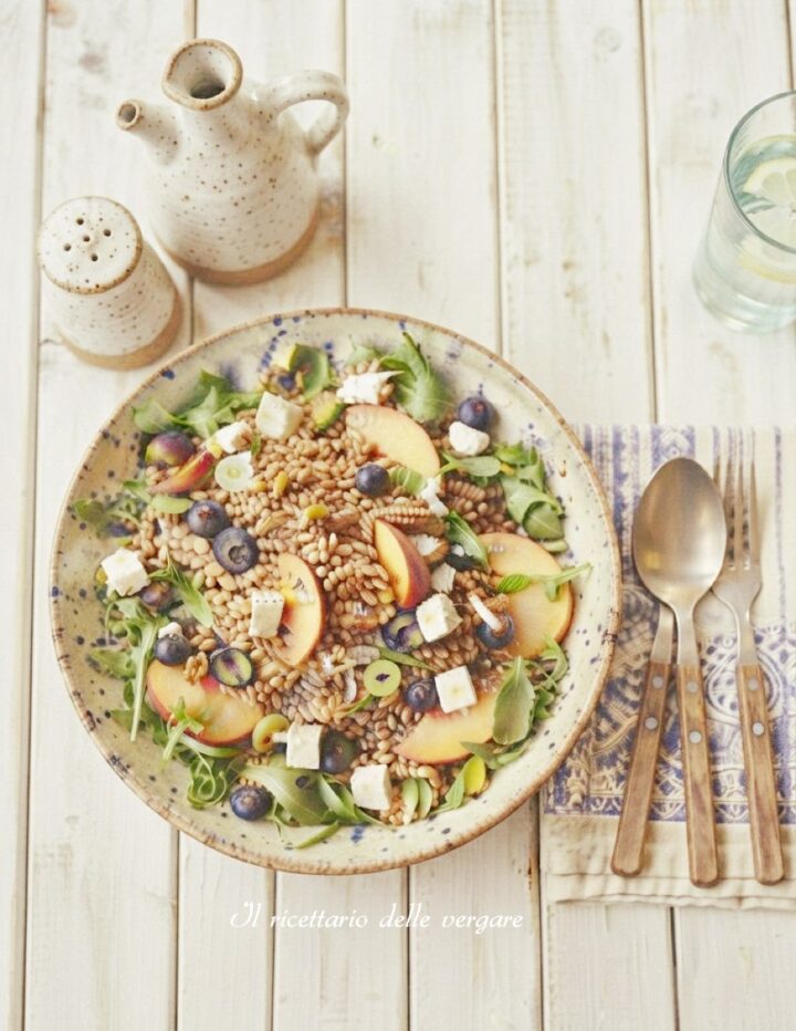 Inquadratura verticale dall'alto di un piatto rustico con insalata di farro estiva, spicchi di pesca, mirtilli, cubetti di formaggio e rucola.