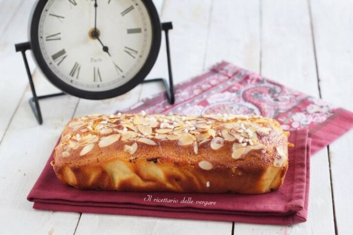Plumcake alla robiola, cocco e mandorle soffice per la colazione, adagiato su un tovagliolo rosso accanto a un orologio antico