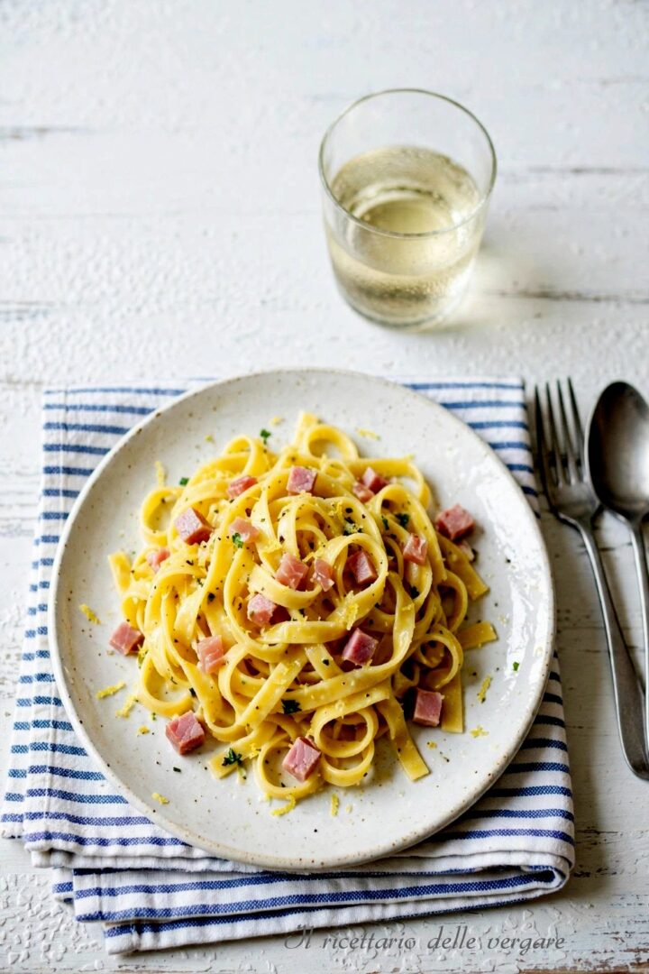 Un primo piano ravvicinato di un piatto di tagliatelle cremose al limone con cubetti di prosciutto cotto, pepe nero e scorza grattugiata.