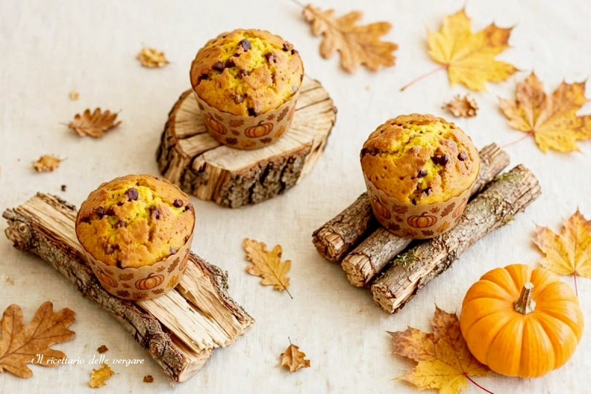 re muffin alla zucca e gocce di cioccolato posizionati su tronchetti di legno decorativi, circondati da foglie autunnali e una piccola zucca arancione