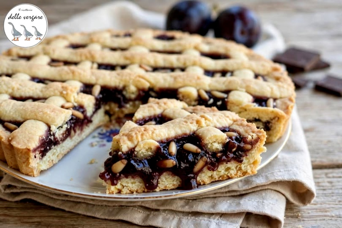 Fetta di crostata con prugne fresche, cioccolato e pinoli su un piatto con prugne e cioccolato sullo sfondo