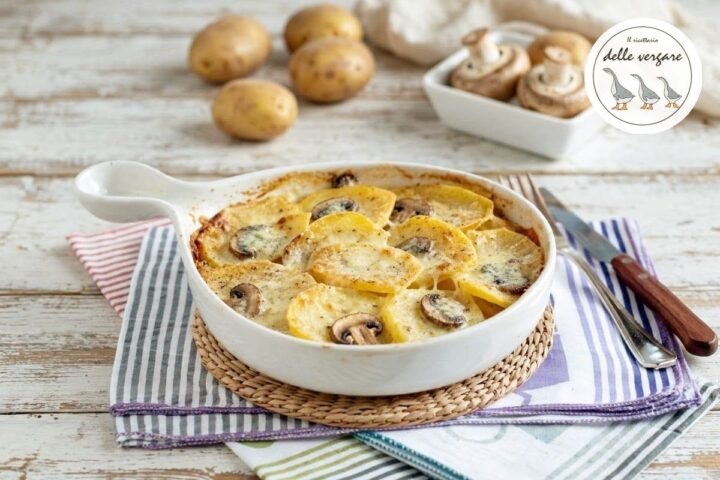 Una pirofila in ceramica bianca con gratin di patate a fette, funghi champignon e gorgonzola fuso, appoggiata su un canovaccio a righe.