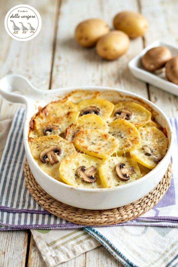 Inquadratura verticale di una pirofila con gratin di patate a fette, funghi champignon e gorgonzola, con patate intere e funghi sullo sfondo.