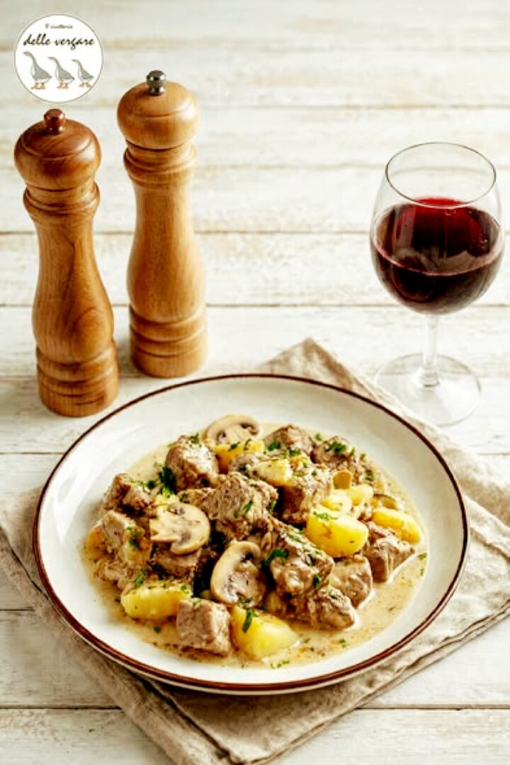 Inquadratura verticale dall'alto di un piatto di spezzatino di vitello in bianco con funghi e patate, accompagnato da un calice di vino rosso e macinapepe in legno.