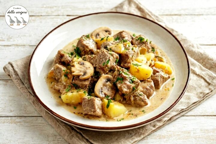 Spezzatino di vitello in bianco con funghi champignon e patate a tocchetti, servito in un piatto fondo decorato con prezzemolo tritato.