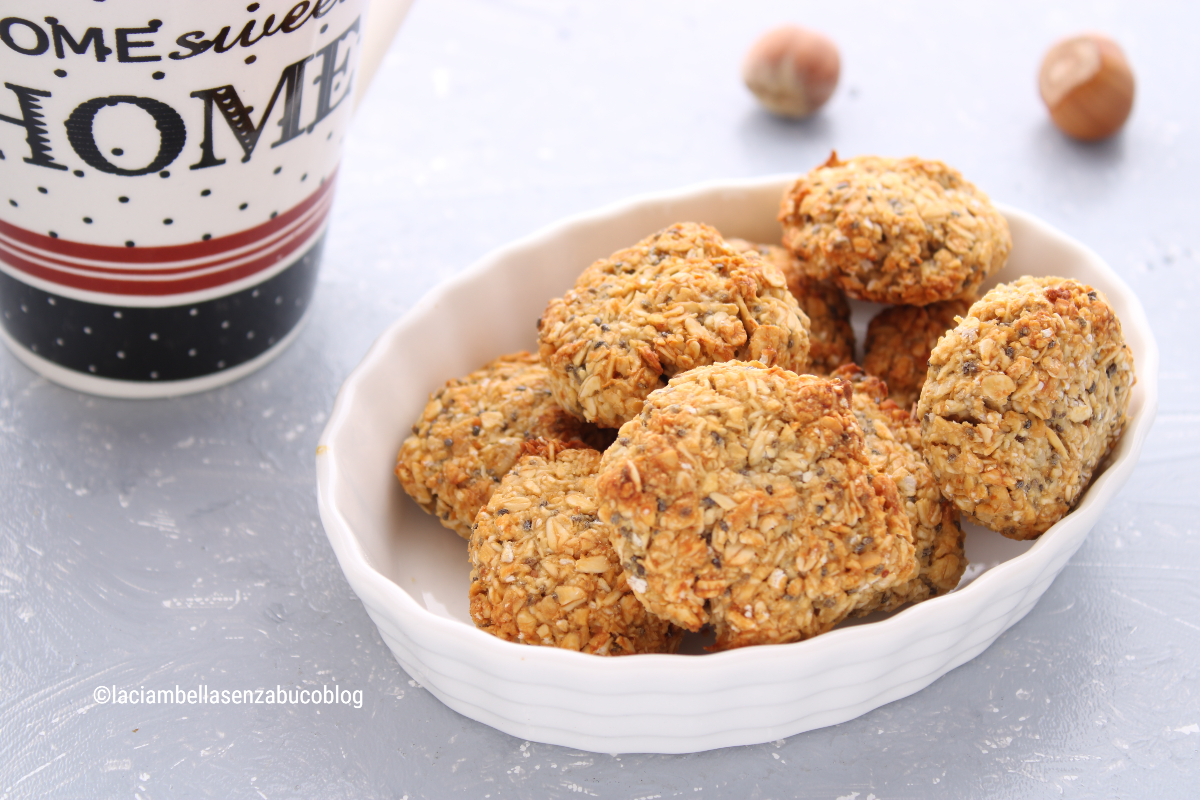 biscotti fiocchi d'avena disposti in un vassoio. Sul piano ci sono 2 nocciole e una tazza da latte.