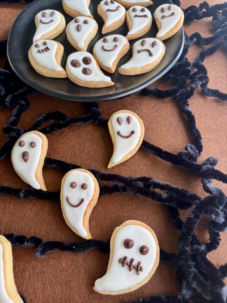Biscotti fantasma di Halloween decorati con ghiaccia bianca e faccine di cioccolato, disposti su un piatto nero.