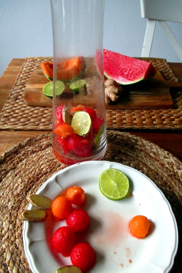 Preparazione bevanda depurativa anguria e melone: frutta a cubetti, lime e zenzero inseriti sul fondo di una caraffa di vetro alta