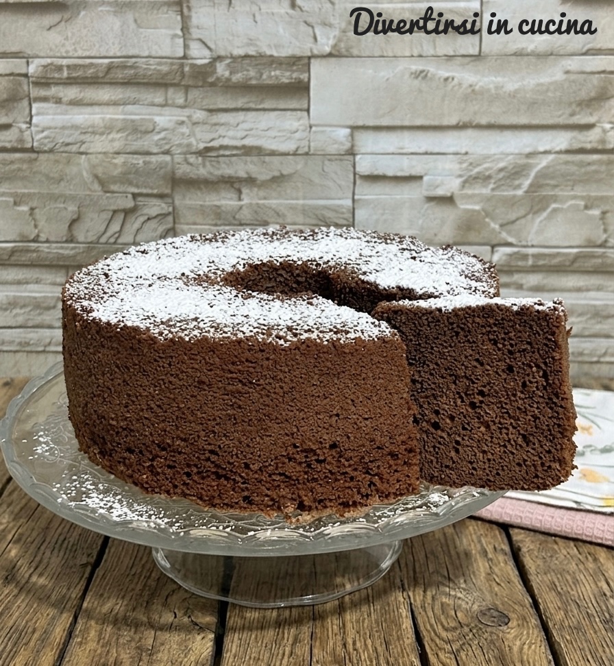 Una fetta generosa di chiffon cake al cacao, tagliata e posizionata a fianco dell'intera torta e ricoperta di zucchero a velo su un'alzatine di vetro e un tavolo in legno rustico.