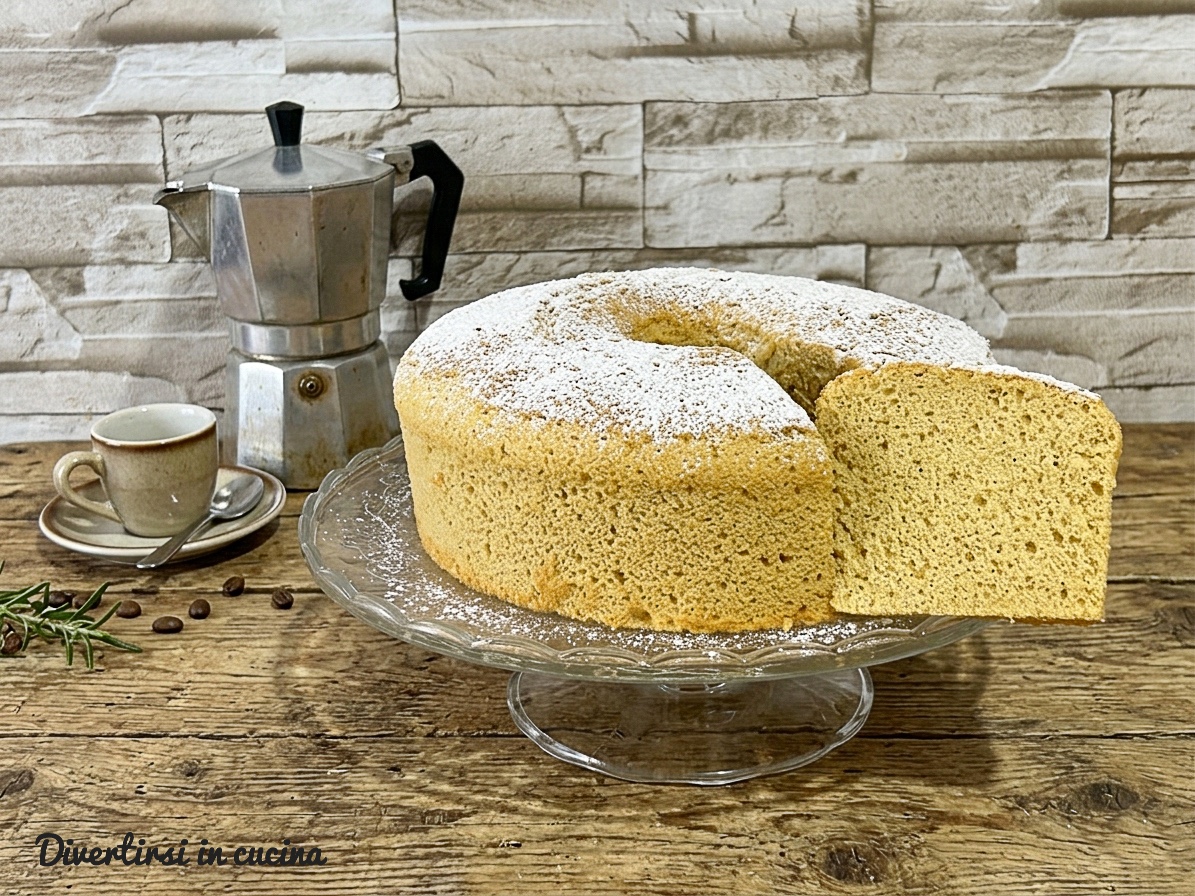 Una soffice e alta chiffon cake al caffè su un'elegante alzatine di vetro, coperta da zucchero a velo. E' stata tagliata una fetta, che mostra l'interno areato e soffice del dolce, posizionata accanto alla torta principale.