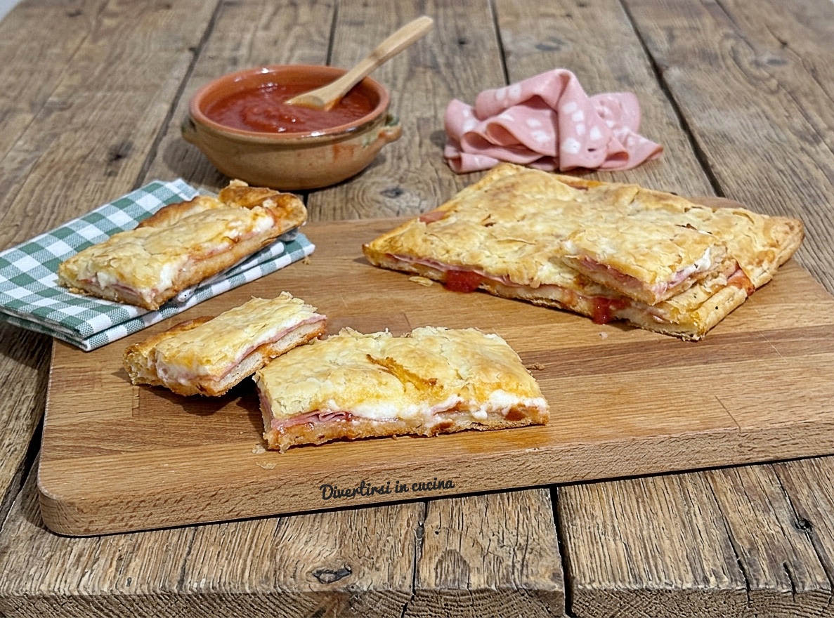 Un tagliere di legno con porzioni tagliate di rustico sfogliato con stracchino e mortadella, affiancate da una ciotolina di salsa di pomodoro e fette di salume, su un tavolo di legno rustico