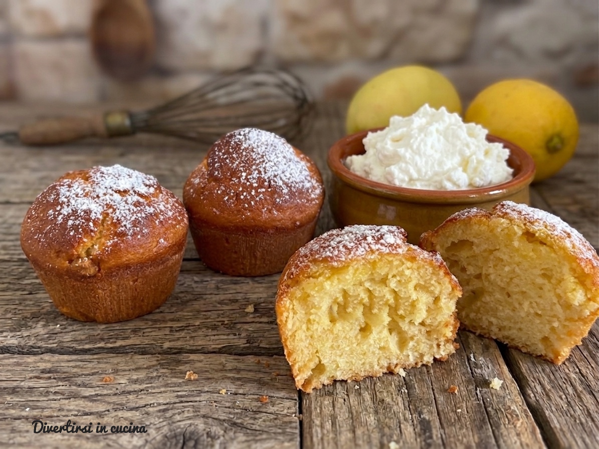 Muffin soffici allo yogurt e limone spolverati di zucchero a velo su un asse di legno rustico con una ciotola di ricotta fresca sullo sfondo