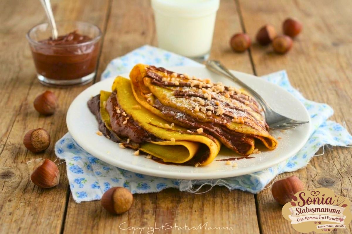 Crepes piegate farcite con crema di nocciole e granella, servite su un piatto con un bicchiere di latte e nocciole intere su un tavolo di legno.